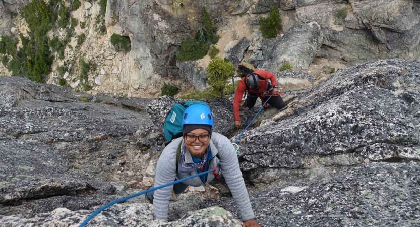 a person looks up and smiles at the camera while rock climbing. another person climbs below her. 